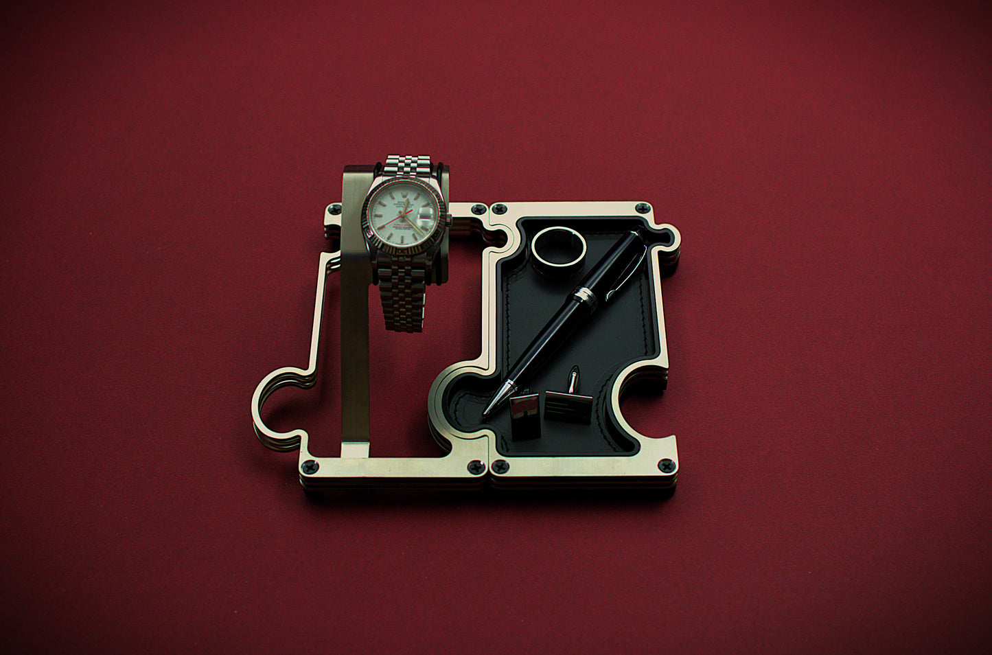Watch and pen on a mechanical component against a red background. The product shown is a combination set of a watch display on the left interlinked with a valet tray on the right. The finish of this product is brushed steel with a silver colour. The watch shown on the watch holder is a Rolex Turn-O-Graph witha white dial and red second hand. The vanity tray to the right shows a leather insert in black real leather with a silver ring, silver cufflinks and a blakc and silver ball point pen